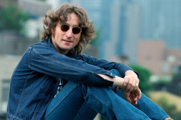 John Lennon com óculos escuros, sentando e com as mãos no joelho