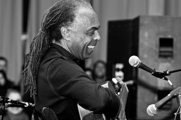 Gilberto Gil com cabelo comprido tocando violão e cantando